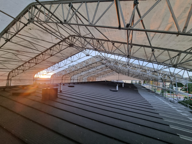Angled view of a building’s dark metal roof beneath a temporary weather-protection canopy, with metal truss framing and translucent sheeting overhead, lit by low sunlight
